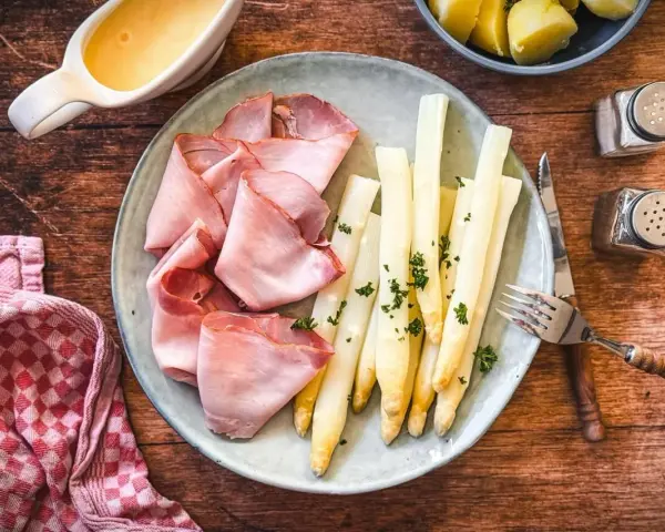 Spargel mit Schinken, garniert mit Petersilie, auf einem Teller mit Sauce Hollandaise und Kartoffeln daneben.