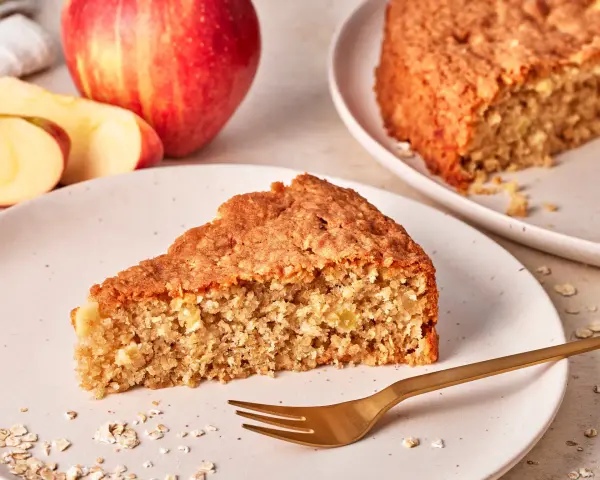 Ein saftiges Stück Apfel Haferflocken Kuchen liegt auf einem Teller, daneben ein ganzer Apfel und Haferflocken.