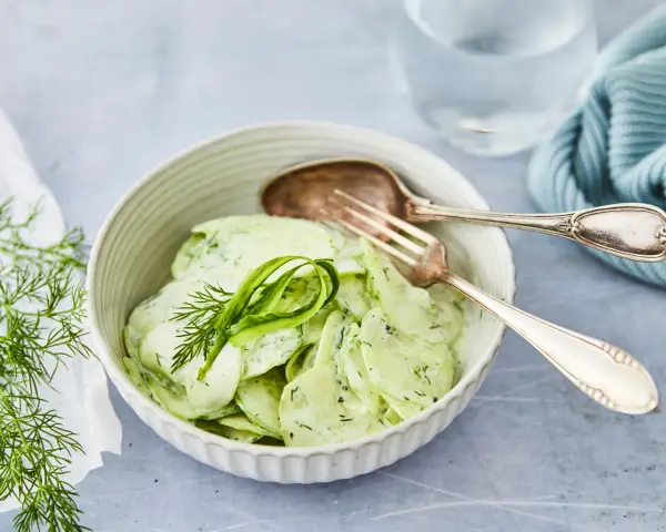 Omas Gurkensalat mit Joghurt, cremig und frisch, garniert mit Dill. Ein Löffel und eine Gabel liegen bereit.