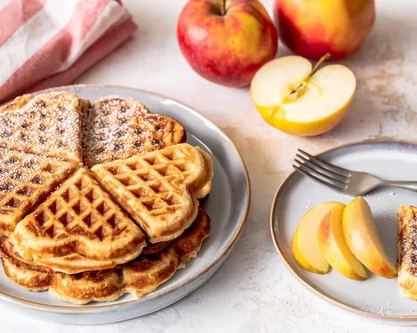 Herzförmige Waffeln mit Puderzucker, daneben frische Äpfel und Apfelspalten. Ein Genuss für alle Sinne!