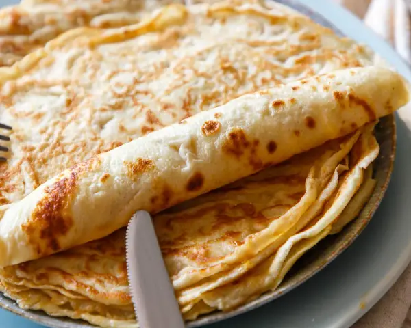 Ein Stapel goldbrauner Pfannkuchen, einer davon aufgerollt, mit Messer und Gabel. Perfekt für ein leckeres Pfannkuchen Rezept!