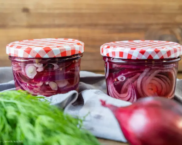 Zwei Gläser mit eingelegten Zwiebeln, rot und weiß, stehen auf einem grauen Tuch. Frischer Dill und eine rote Zwiebel liegen davor.