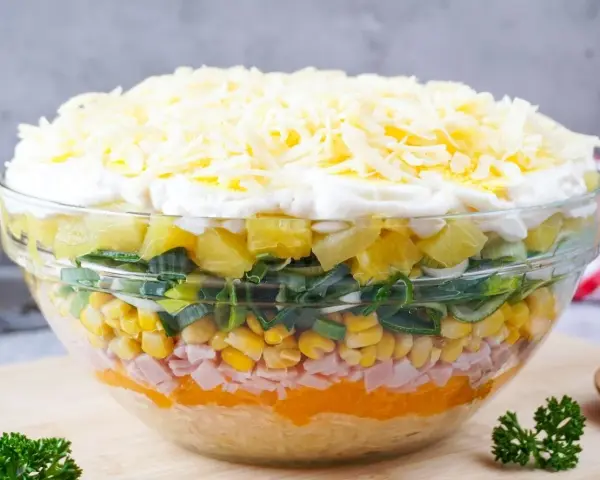 Schichtsalat rezept: Ein bunter Schichtsalat in einer Glasschüssel mit Ananas, Lauch, Mais, Schinken, Orange, Ei und Käse.