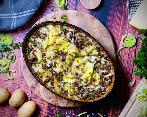 Herzhafter kartoffelauflauf mit hackfleisch, überbacken mit Käse und garniert mit Petersilie. Dazu frische Kartoffeln, Lauch und ein Stück Käse.