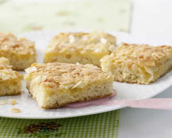 Saftiger mandelkuchen vom blech, goldbraun gebacken und mit knusprigen Mandelblättchen bestreut, liegt auf einer weißen Platte.