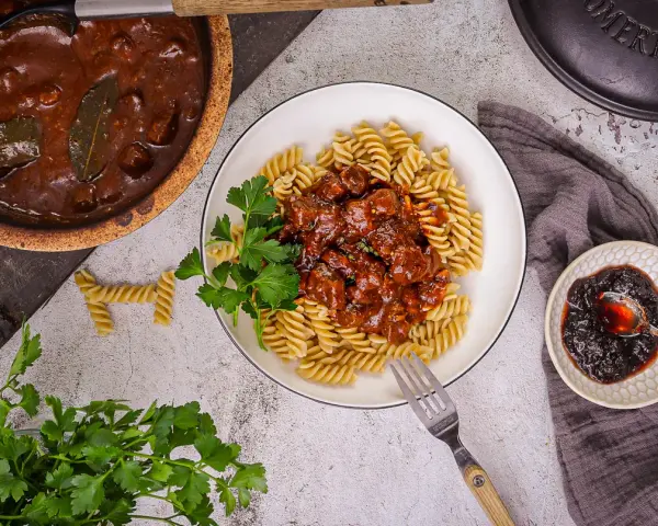 Herzhaftes Gulasch gemischt Rezept mit Fusilli, garniert mit Petersilie. Daneben ein kleiner Teller mit Preiselbeeren.