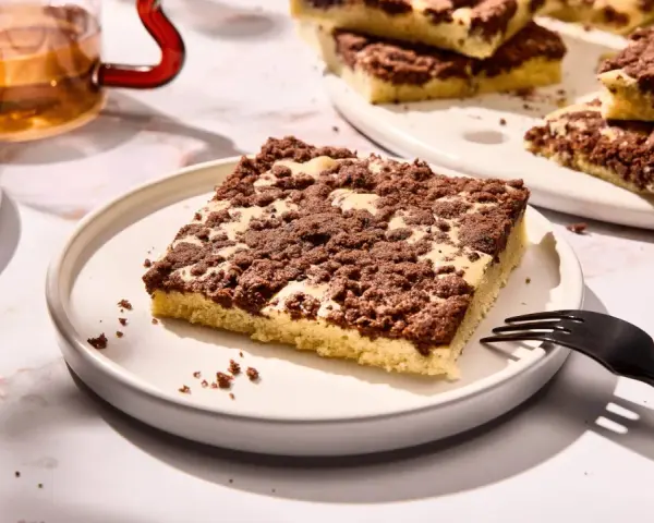 Ein Stück versunkener Streuselkuchen mit Schokostreuseln auf einem Teller, daneben eine Gabel und ein Glas Tee.