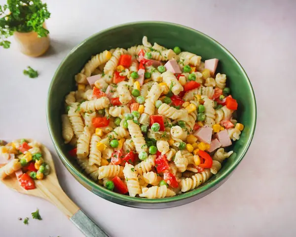 Ein bunter Nudelsalat mit Fleischwurst, Erbsen, Mais und Paprika in einer grünen Schüssel.