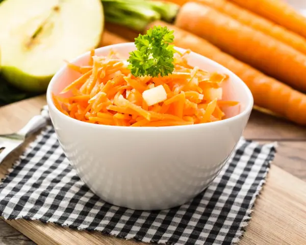 Frischer Karotten-Apfel-Salat in einer weißen Schüssel, garniert mit Petersilie. Im Hintergrund liegen ganze Karotten und ein halber Apfel.