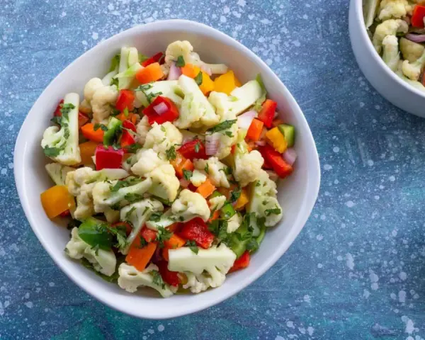 Frischer blumenkohl salat mit Paprika, Karotten und Kräutern, serviert in einer weißen Schüssel. Daneben eine kleine Schale mit Dip.