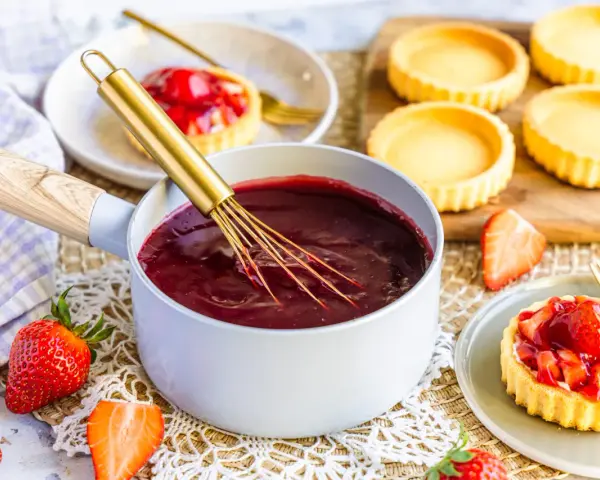 Frisch zubereiteter Tortenguss in einem Topf, bereit, um Obstkuchen zu verfeinern. Daneben liegen frische Erdbeeren und leere Tartelettes.
