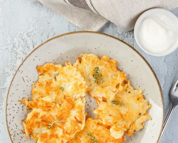 Knusprige Kartoffelpuffer, im Waffeleisen goldbraun gebacken, liegen auf einem Teller. Dazu ein Schälchen mit Sour Cream und ein Gabel.
