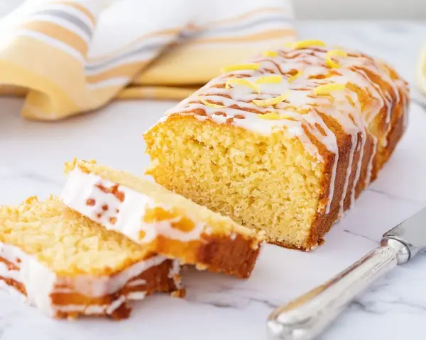 Saftiger zitronenkuchen mit Joghurt, überzogen mit Zuckerguss und garniert mit Zitronenschale. Ein Stück ist angeschnitten.