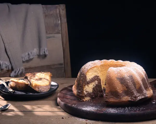 Ein fluffiger Marmorkuchen, bestäubt mit Puderzucker, liegt auf einem Holzbrett. Daneben zwei Stücke auf einem Teller.