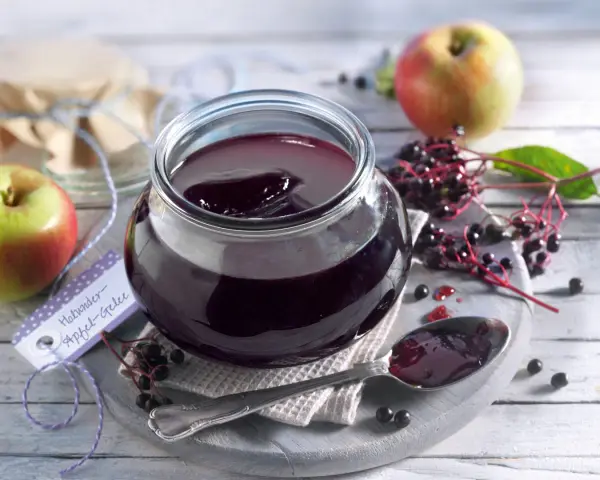 Ein Glas mit dunklem Holunder Apfel Gelee, daneben Äpfel und Holunderbeeren. Ein Löffel mit Gelee liegt bereit.