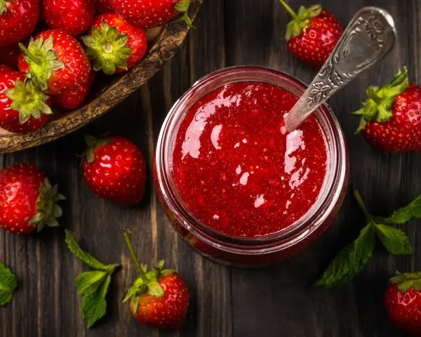 Frische Erdbeeren und ein Glas selbstgemachte Erdbeermarmelade, bereit zum Einkochen. Ein Löffel steckt in der Marmelade.