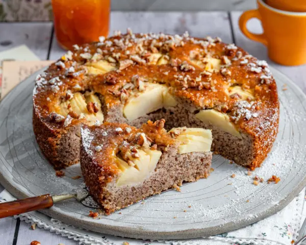 Ein saftiger Apfel-Birnen-Kuchen, bestreut mit Nüssen und Puderzucker, wird auf einem Teller serviert. Ein Stück ist bereits herausgeschnitten.