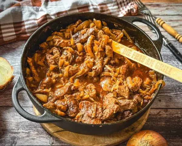 Herzhaftes Zwiebelfleisch-Rezept in Gusseisenpfanne, mit Zwiebeln und Fleischstücken in einer würzigen Sauce. Ein Holzlöffel mit der Aufschrift "Die Frau am Grill" liegt darin.