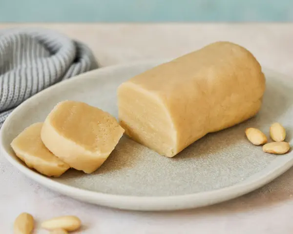 Frisch zubereitetes Marzipan zum **Marzipan selbst machen**. Ein Block und zwei Scheiben liegen auf einem Teller, daneben Mandeln.