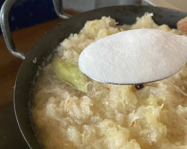 Zucker wird zu köchelndem Sauerkraut nach Omas Rezept gegeben. Ein Lorbeerblatt schwimmt im Topf.