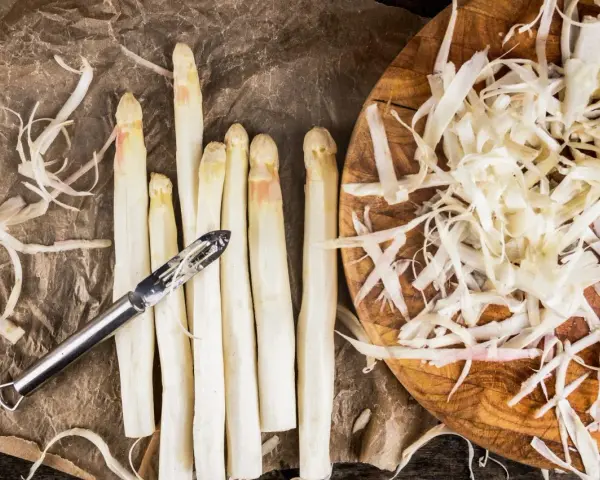 Frischer weißer Spargel wird geschält, die Spargelschalen liegen daneben. Ein Sparschäler liegt auf den Spargelstangen.