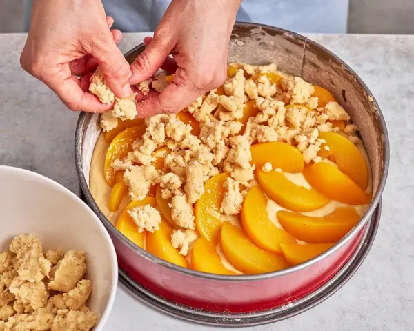 Hände verteilen Streusel auf einem Pfirsichkuchen in einer Springform.