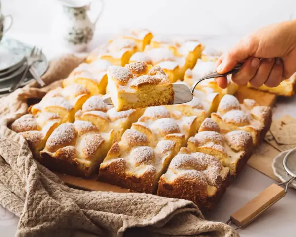 Ein Stück Spiegeleikuchen mit Quark wird aus einem Blechkuchen mit Puderzucker gehoben.