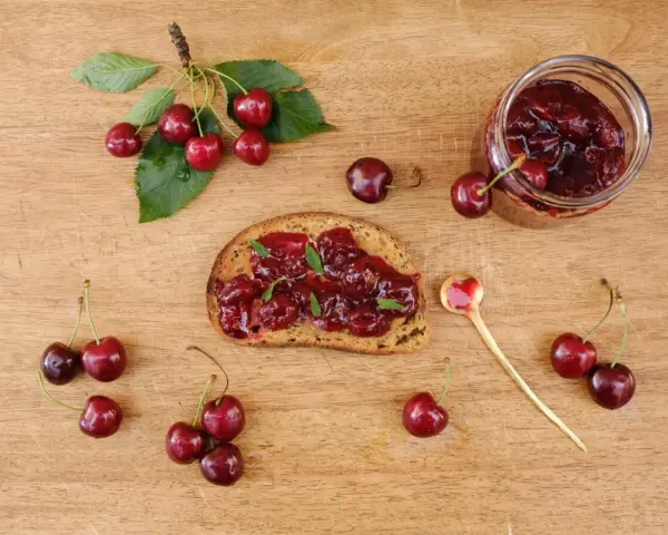 Frisches Brot mit Kirschmarmelade, garniert mit Minze. Ein Glas Kirschmarmelade und frische Kirschen daneben. Perfekt für ein Kirschmarmelade Rezept.
