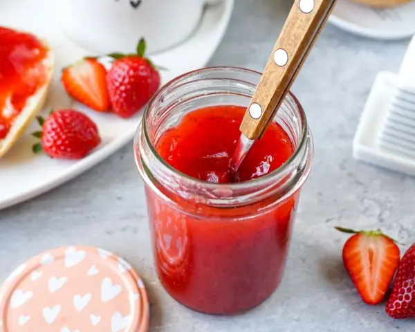 Ein Glas mit leuchtend roter Erdbeermarmelade, die mit einem Holzlöffel umgerührt wird. Daneben frische Erdbeeren und ein Stück Brot mit Marmelade – ein Genuss mit Pfiff!