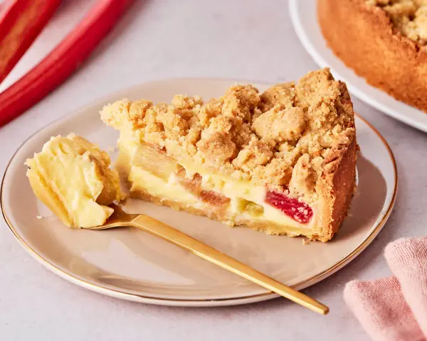 Ein Stück von Omas Rhabarberkuchen mit Pudding und Streuseln auf einem Teller, daneben ein goldener Löffel und rote Rhabarberstangen.