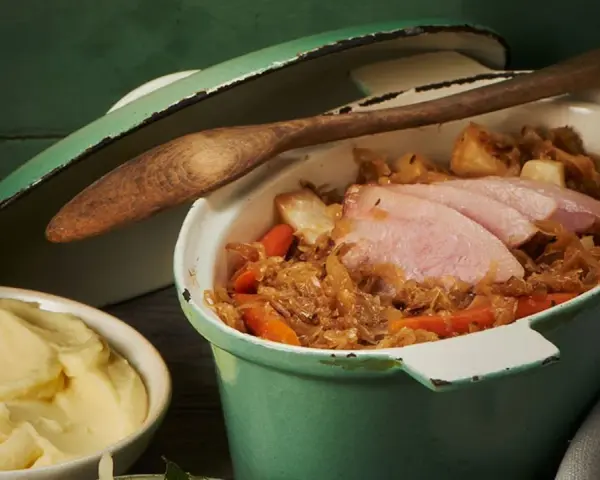 Herzhafter Szegediner Gulasch mit zarten Fleischscheiben, Sauerkraut und Karotten in einem grünen Topf, daneben eine Schüssel cremiges Kartoffelpüree.