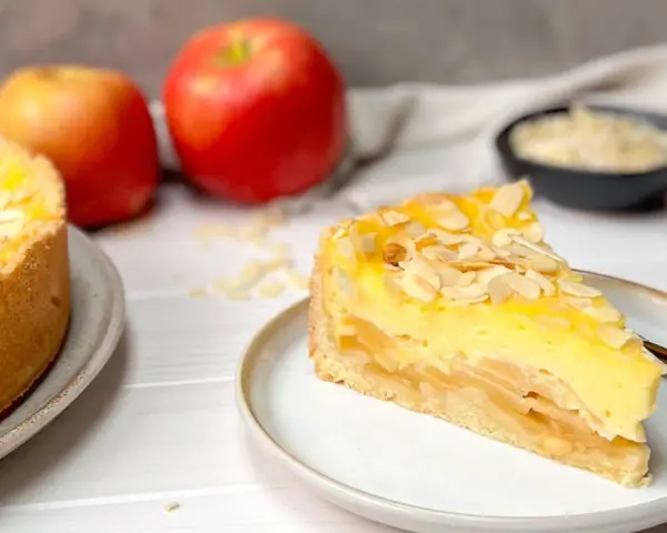 Ein saftiger Apfelkuchen mit Schmandguss, bestreut mit Mandelblättchen. Daneben liegen frische Äpfel.