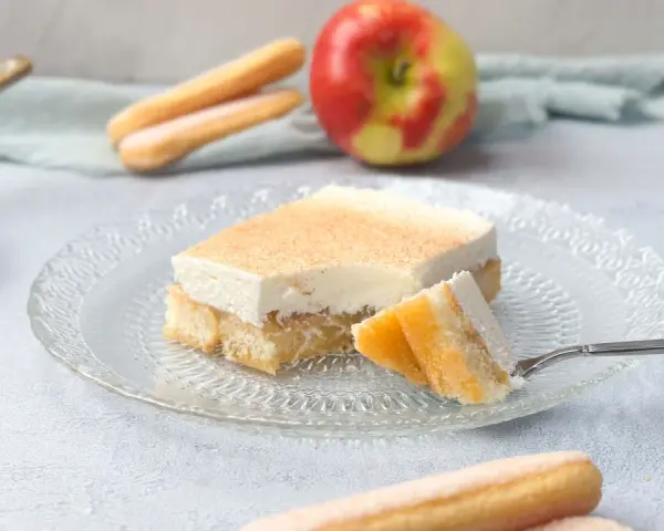 Ein Stück Apfel-Schmand-Kuchen aus der Springform, mit Löffelbiskuits und einem Apfel im Hintergrund.