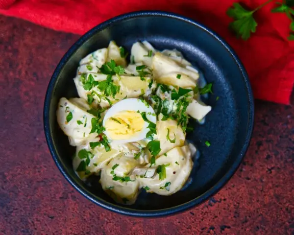 Ein cremiger Kartoffelsalat mit Mayo, garniert mit Petersilie und einem halben Ei, serviert in einer dunklen Schüssel.