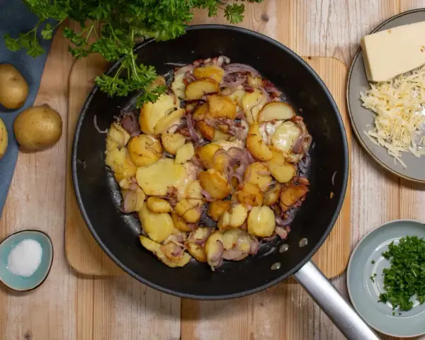 Bratkartoffeln mit Zwiebeln und Käse in der Pfanne, dazu frische Petersilie und Salz.