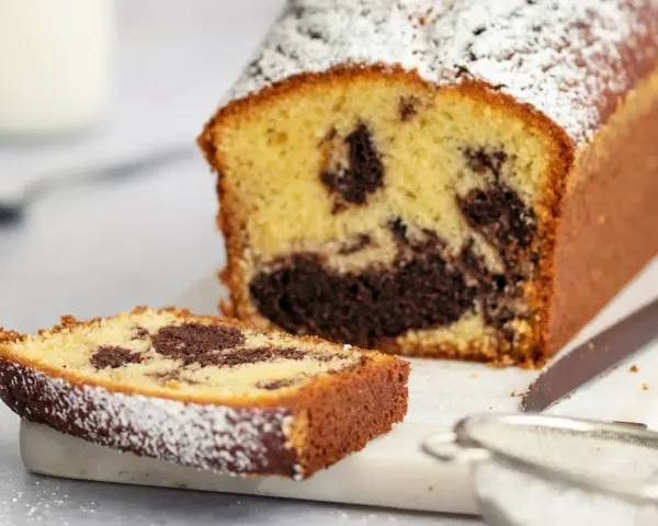 Ein saftiger marmorkuchen mit Puderzucker bestäubt, daneben ein Stück Kuchen und ein Sieb. Perfekt mit einem Klecks Sahne.