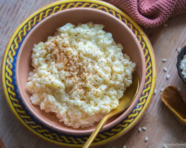 Cremiger Milchreis, bestreut mit Zimt, wartet in einer Schale auf sein Milchreis Rezept. Daneben liegen Reiskörner und Zimtstangen.