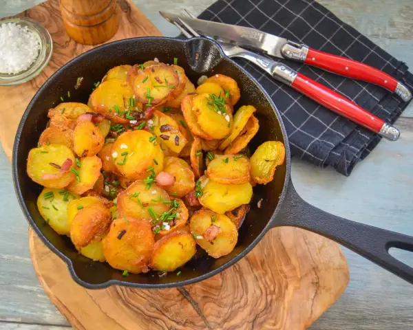 Bratkartoffeln mit Zwiebeln und Speck in einer Gusseisenpfanne, garniert mit Schnittlauch. Dazu grobes Salz und ein Messer-Gabel-Set.