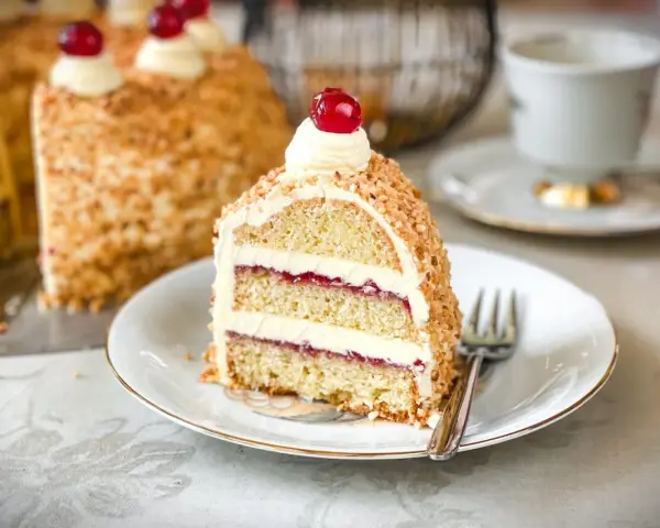 Ein Stück Frankfurter Kranz, das Rezept eines Konditors, mit Buttercreme und Kirschen verziert, umgeben von gehackten Nüssen.