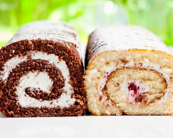 Zwei Biskuitrollen, eine schokoladig, die andere hell, mit Sahne und Fruchtfüllung, bestäubt mit Puderzucker. Wie von Landfrauen gebacken.