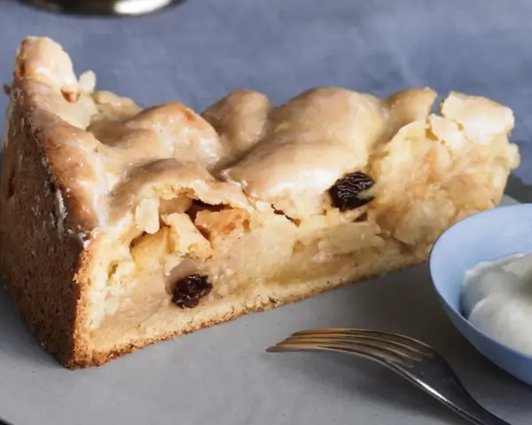 Ein Stück gedeckter Apfelkuchen mit Rosinen und Zuckerguss, serviert mit einem Klecks Sahne.