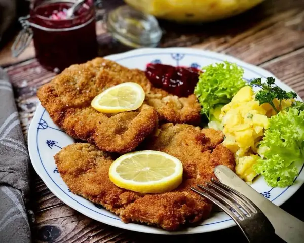Knuspriges Wiener Schnitzel mit Zitrone, Preiselbeeren und Kartoffelsalat auf einem Teller.