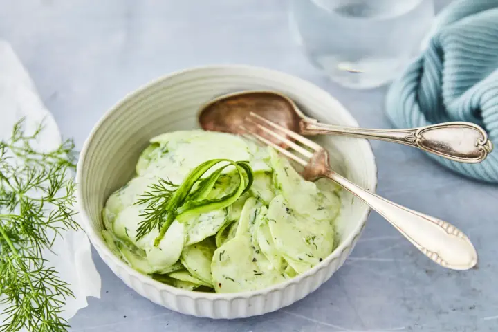 Omas Gurkensalat mit Joghurt, cremig und frisch, garniert mit Dill. Ein Löffel und eine Gabel liegen bereit.