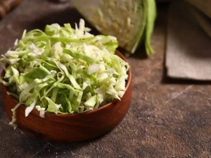 Fein geschnittener Spitzkohl in einer Holzschale, bereit, in der Pfanne gebraten zu werden. Ein halber Spitzkohl liegt im Hintergrund.
