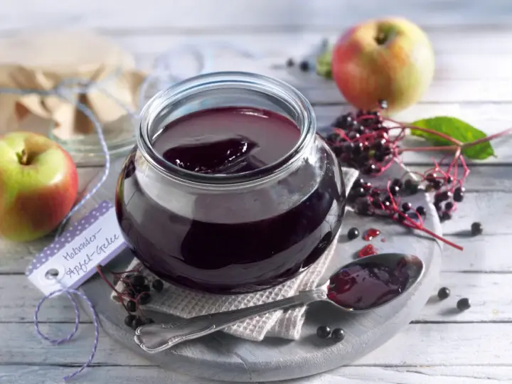 Ein Glas mit dunklem Holunder Apfel Gelee, daneben Äpfel und Holunderbeeren. Ein Löffel mit Gelee liegt bereit.