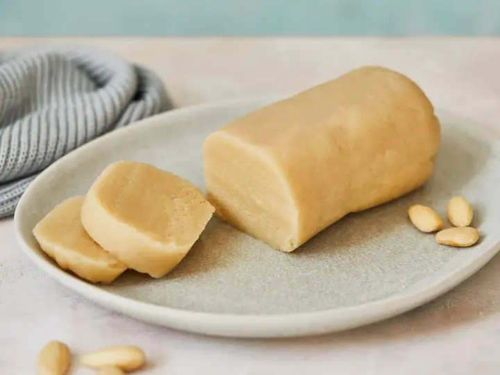 Frisch zubereitetes Marzipan zum **Marzipan selbst machen**. Ein Block und zwei Scheiben liegen auf einem Teller, daneben Mandeln.