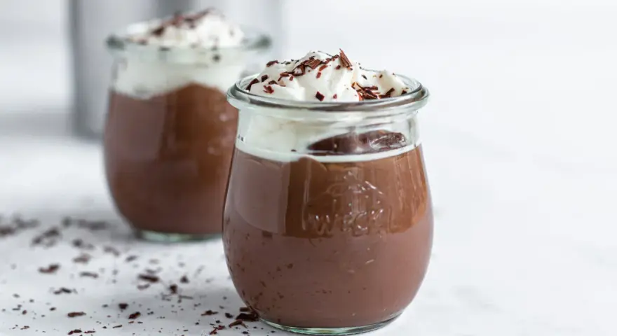 Zwei Gläser mit cremigem Schokopudding, gekrönt mit Schlagsahne und Schokostreuseln. Perfekt, um Schokopudding selber machen zu lernen!
