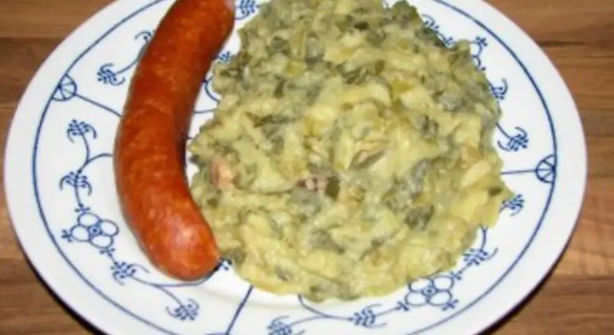 Omas Stielmus Rezept: deftiger Grünkohl mit Kartoffeln und einer herzhaften Bratwurst auf einem blau-weißen Teller.