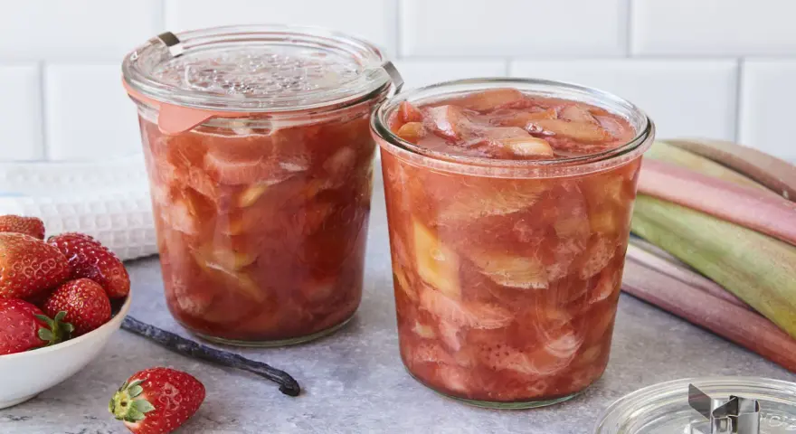 Zwei Gläser mit leckerem Rhabarberkompott, daneben frische Erdbeeren und Rhabarberstangen.