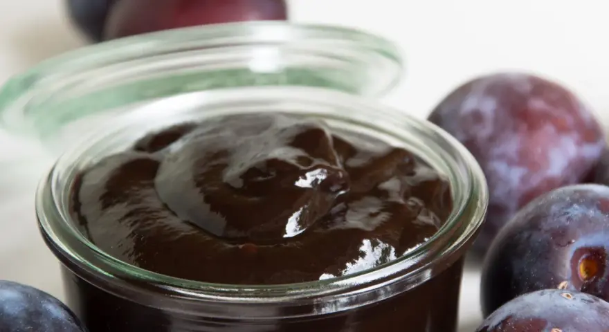 Ein Glas mit dunklem Pflaumenmus, umgeben von frischen Pflaumen. Perfekt für ein einfaches Pflaumenmus Rezept.
